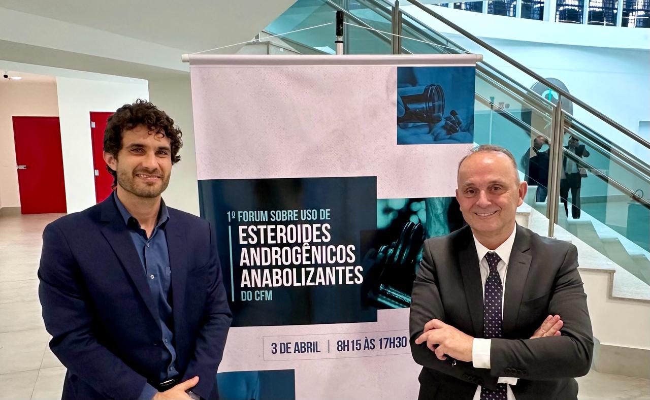 Dois médicos posando sorridentes ao lado do banner do "1º Fórum sobre Uso de Esteroides Androgênicos Anabolizantes do CFM".